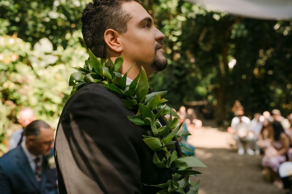 Groom wearing a green leaf lei at an outdoor wedding ceremony surrounded by lush trees and guests.