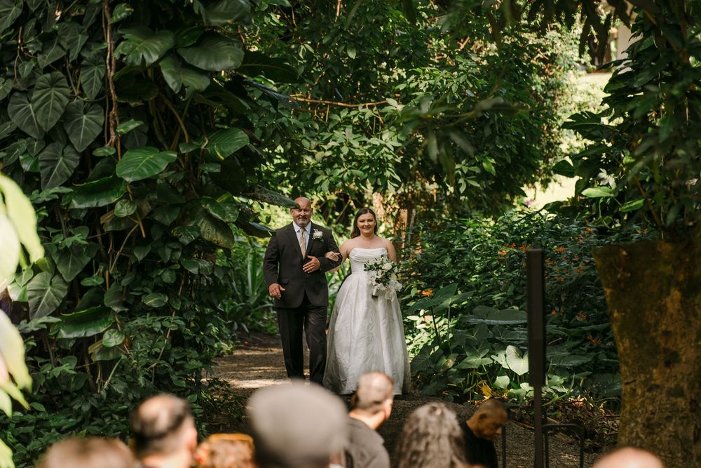 Bride and groom walking down a lush garden pathway surrounded by tropical greenery during an outdoor wedding ceremony.