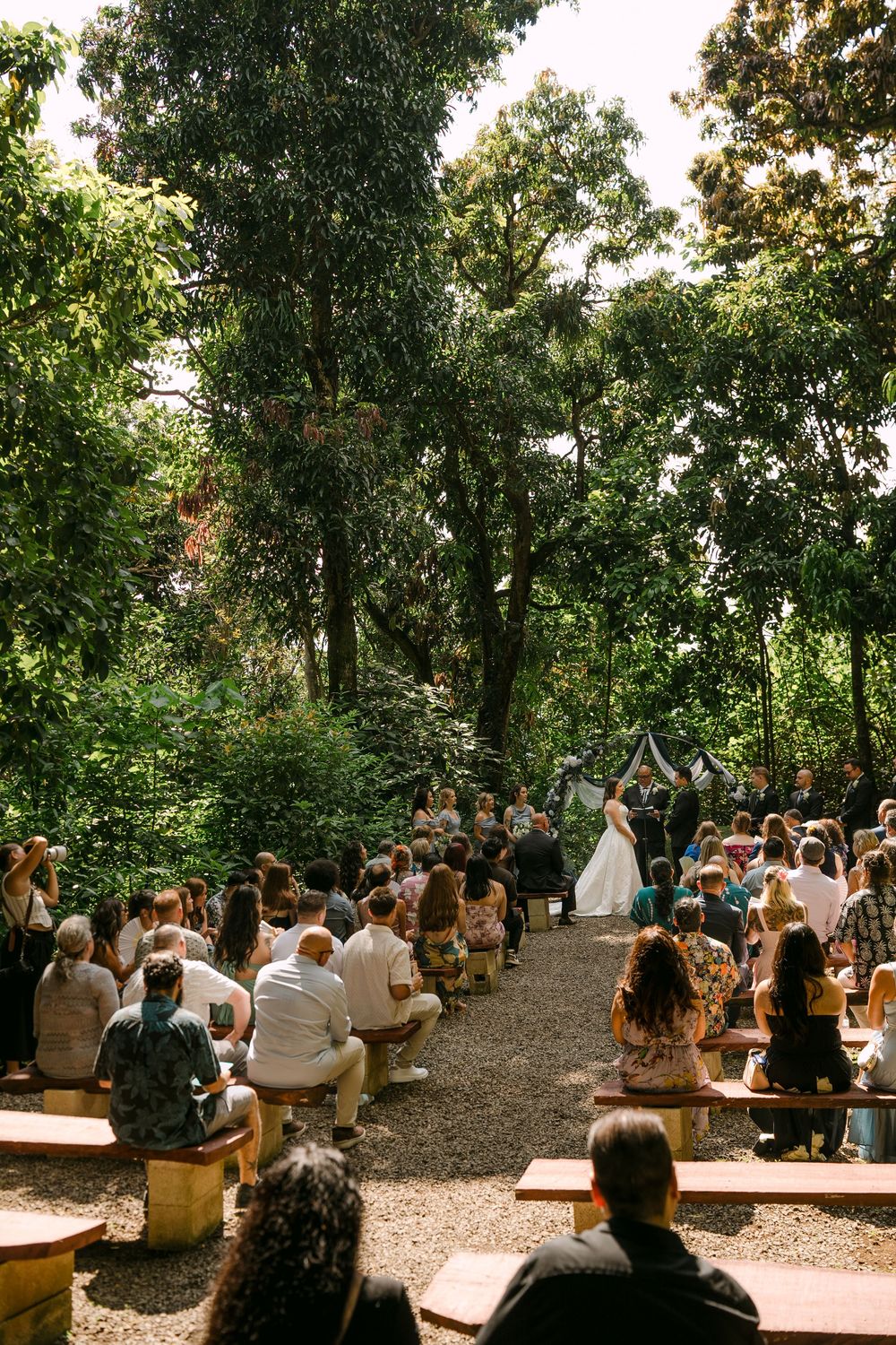 Outdoor wedding ceremony in a lush forest setting with guests seated on wooden benches as bride stands at the altar.