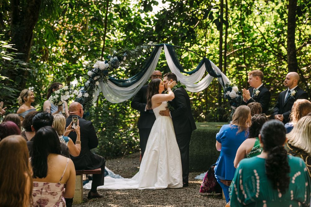 Bride and groom share first kiss at outdoor woodland wedding ceremony with floral arch and seated guests.