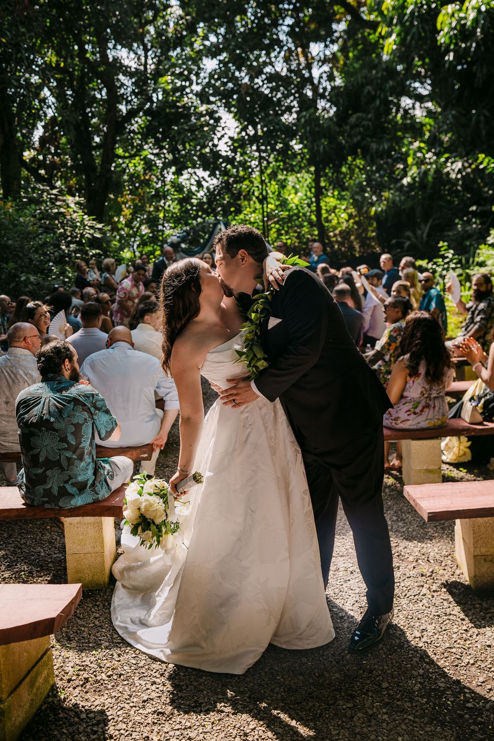 Bride and groom share first kiss as newlyweds in an outdoor garden ceremony aisle, surrounded by seated guests.