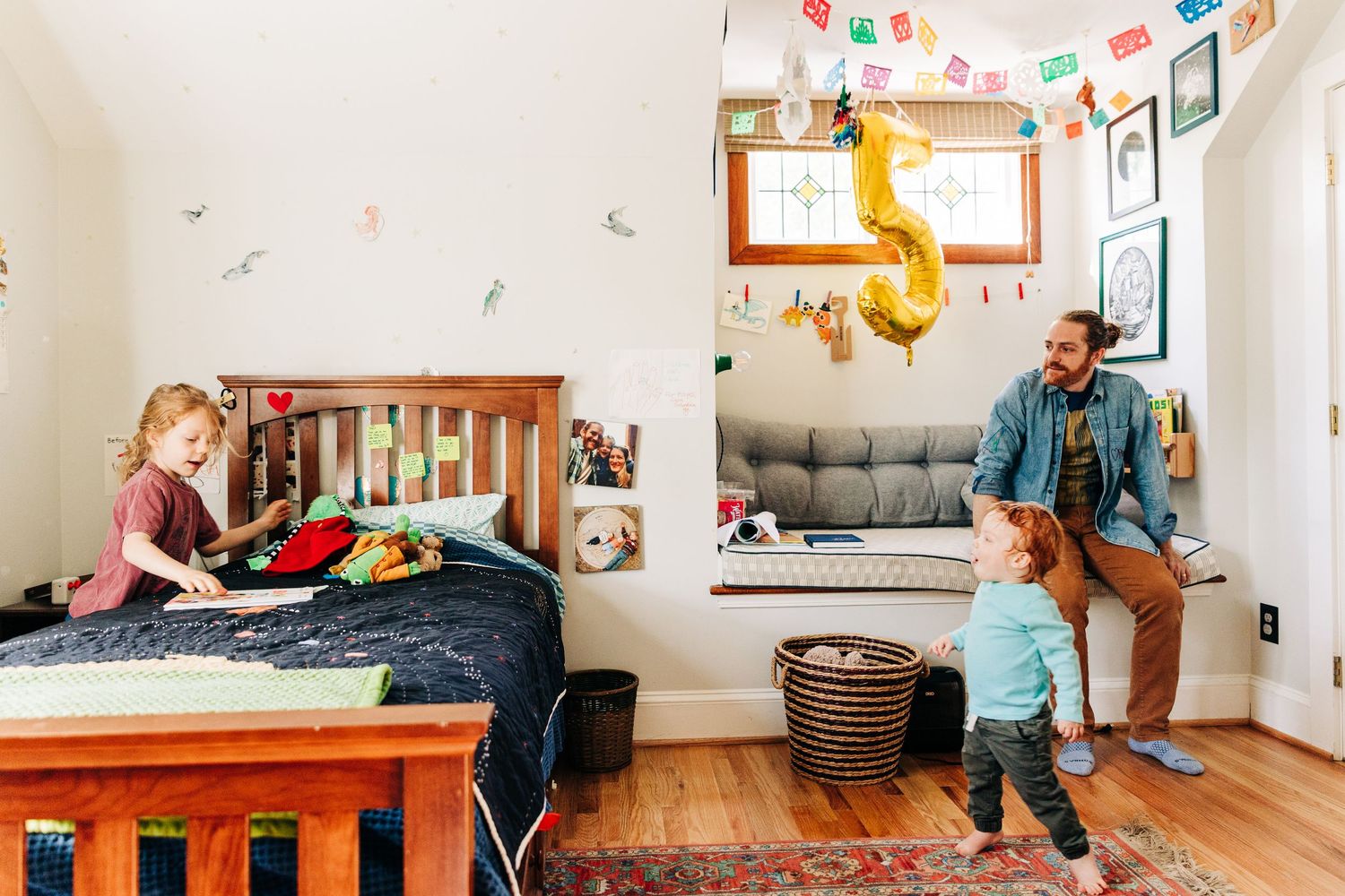 A father and two young children play in a colorful, decorated kids bedroom with a wooden bed and gallery wall in Charlotte, NC.