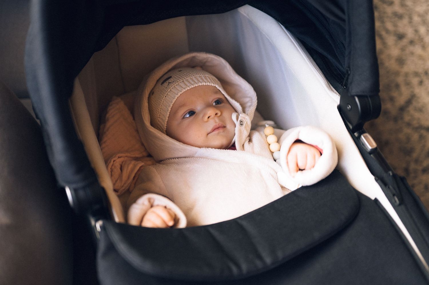 A newborn baby wearing a pink knit hat lies snugly wrapped in a cozy stroller bassinet.