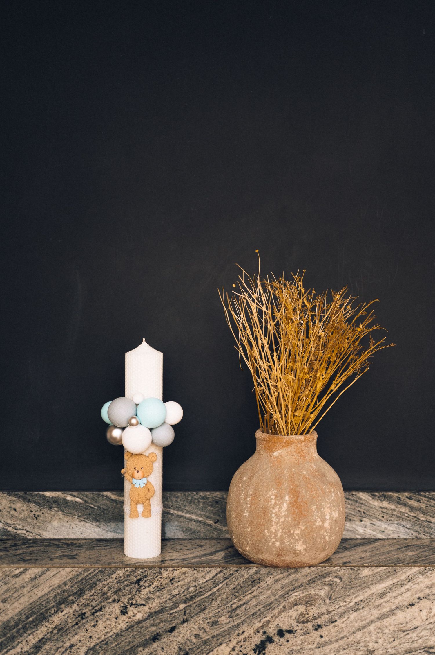 White pillar candle with teddy bear and blue baubles beside a rustic terracotta vase with dried golden branches.