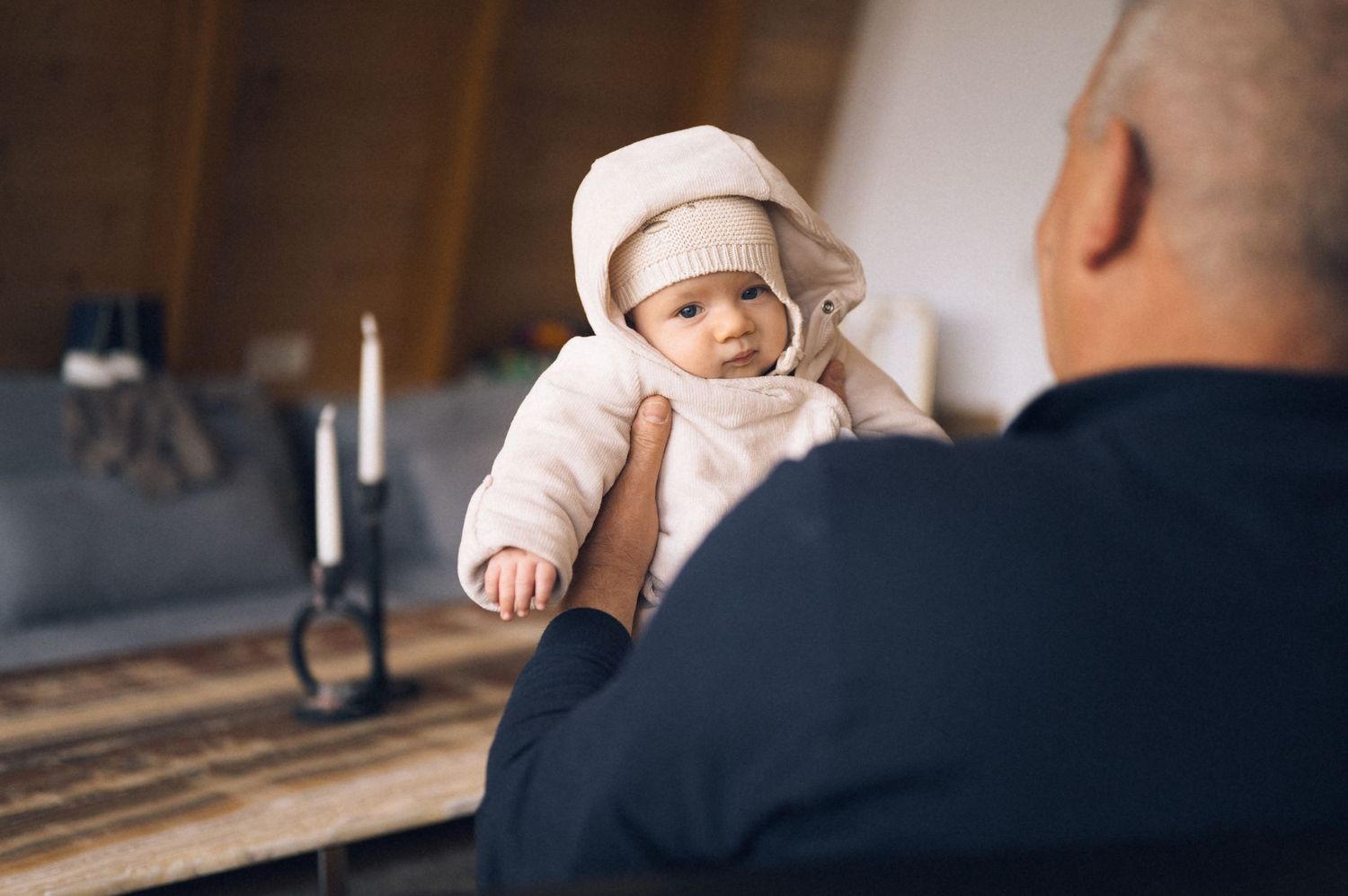 Elderly man holding a cute baby dressed in a cozy white knit hat and jacket indoors.