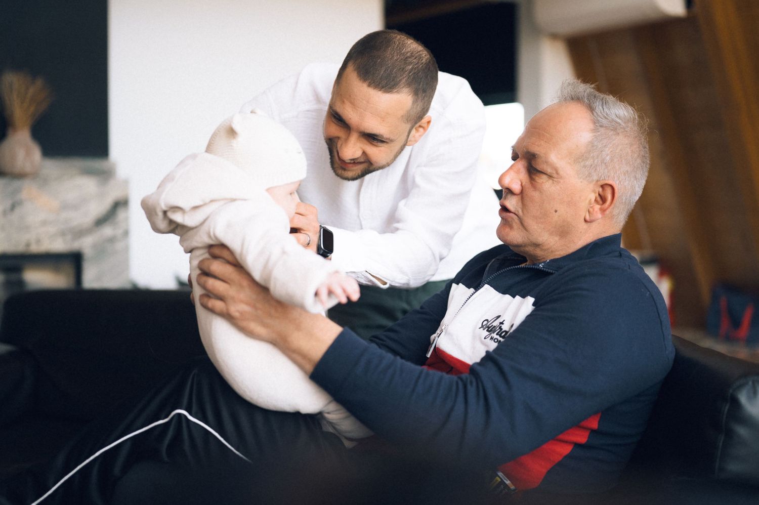 Two men sharing a tender moment with a newborn baby wrapped in white blanket indoors.