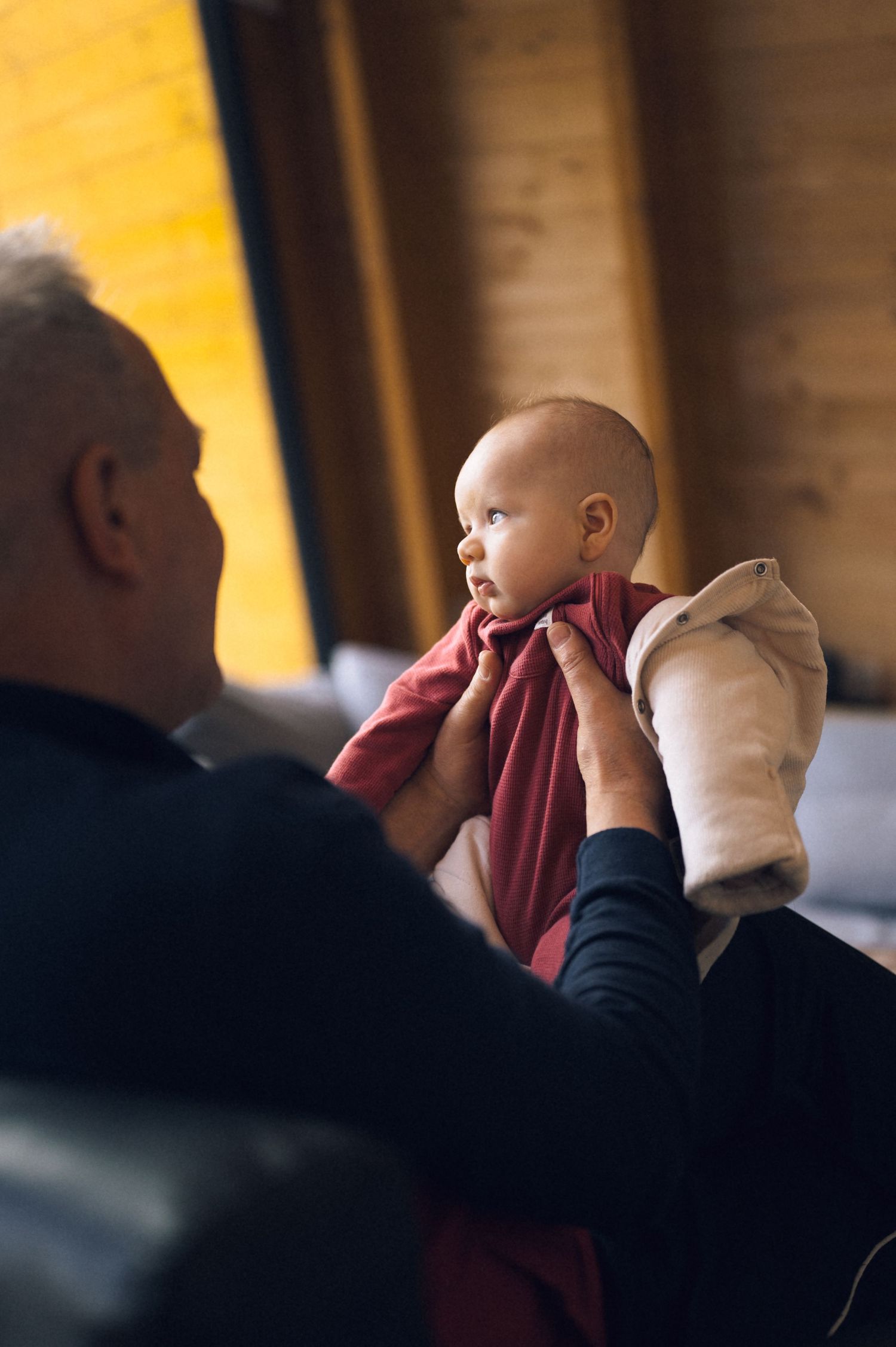 Elderly man holding a newborn baby wrapped in a red blanket while sitting on a couch indoors.