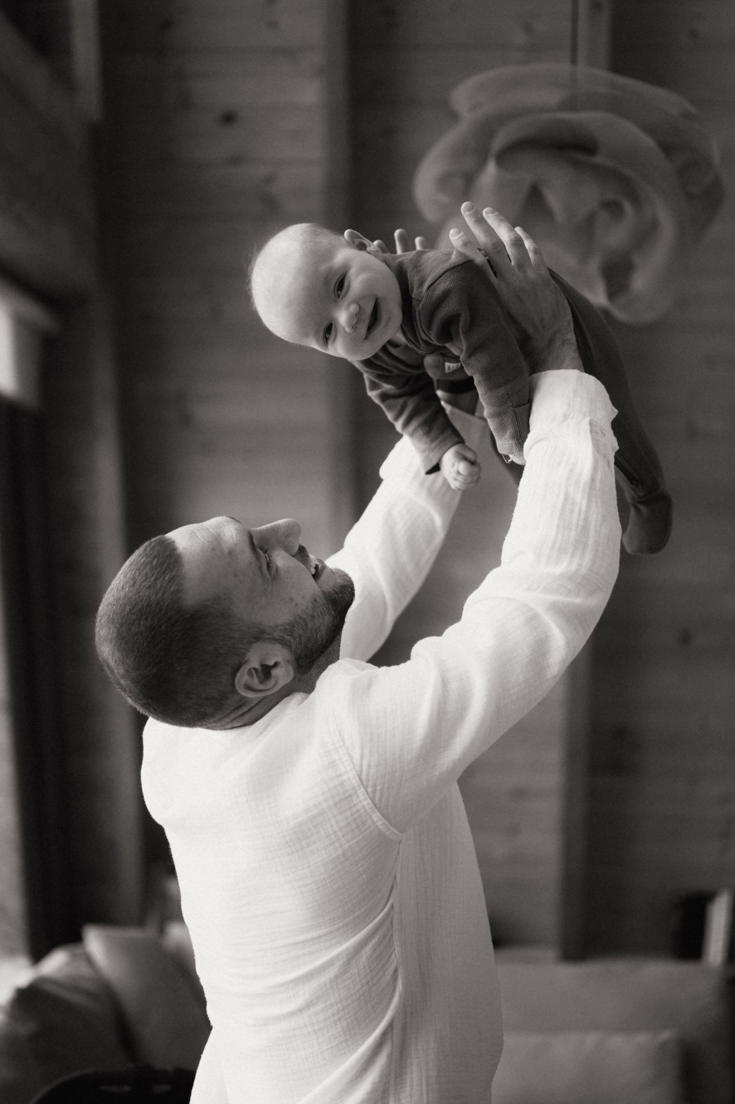Black and white photo of a father lifting his smiling baby overhead indoors.