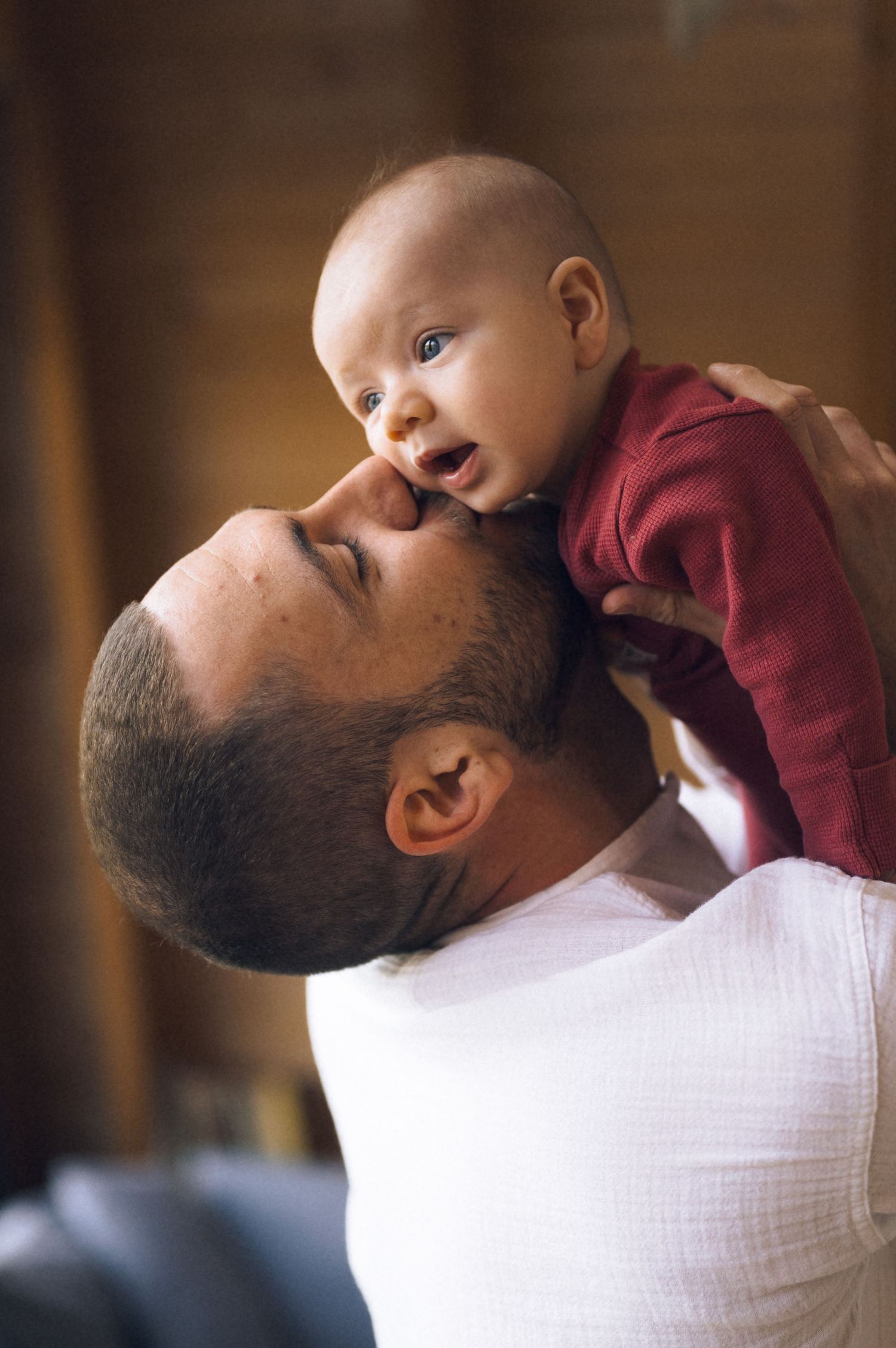 Father lovingly holds smiling baby in red outfit close to his face indoors in warm light.
