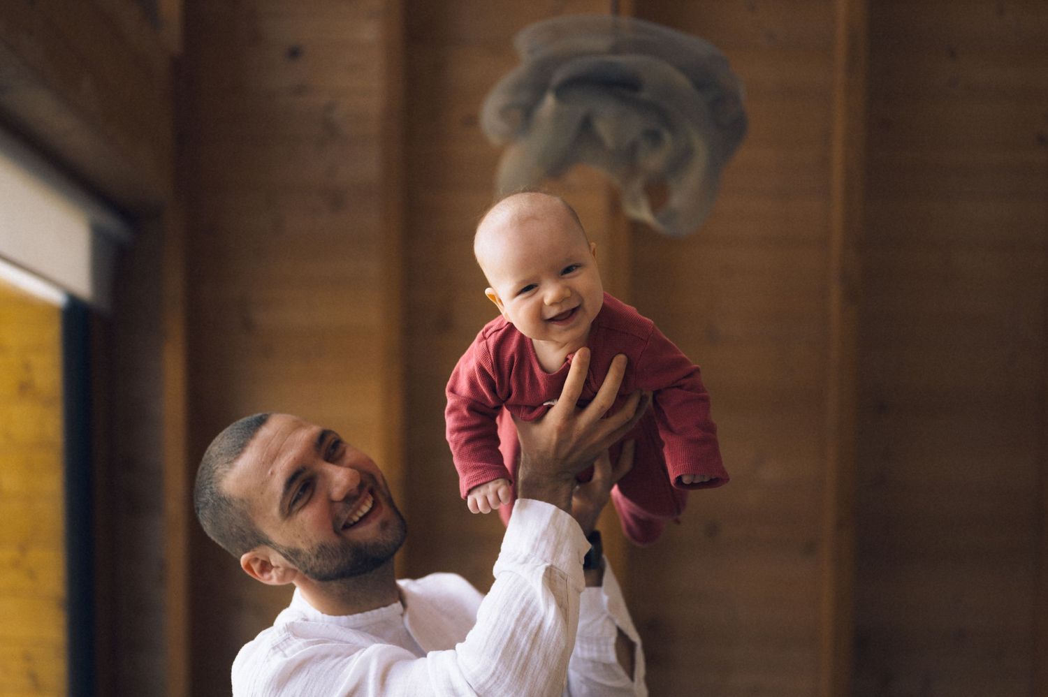 Joyful father lifting laughing baby in red outfit overhead inside a warmly lit wooden interior.