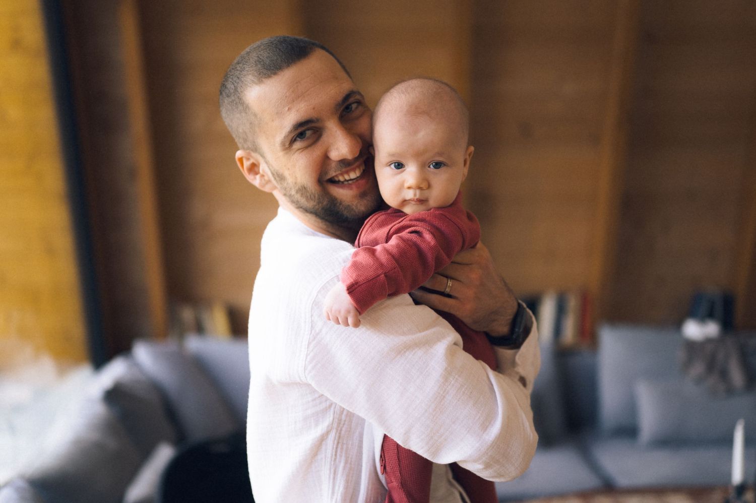 Smiling father holding his newborn baby in a cozy living room, wearing a white shirt.
