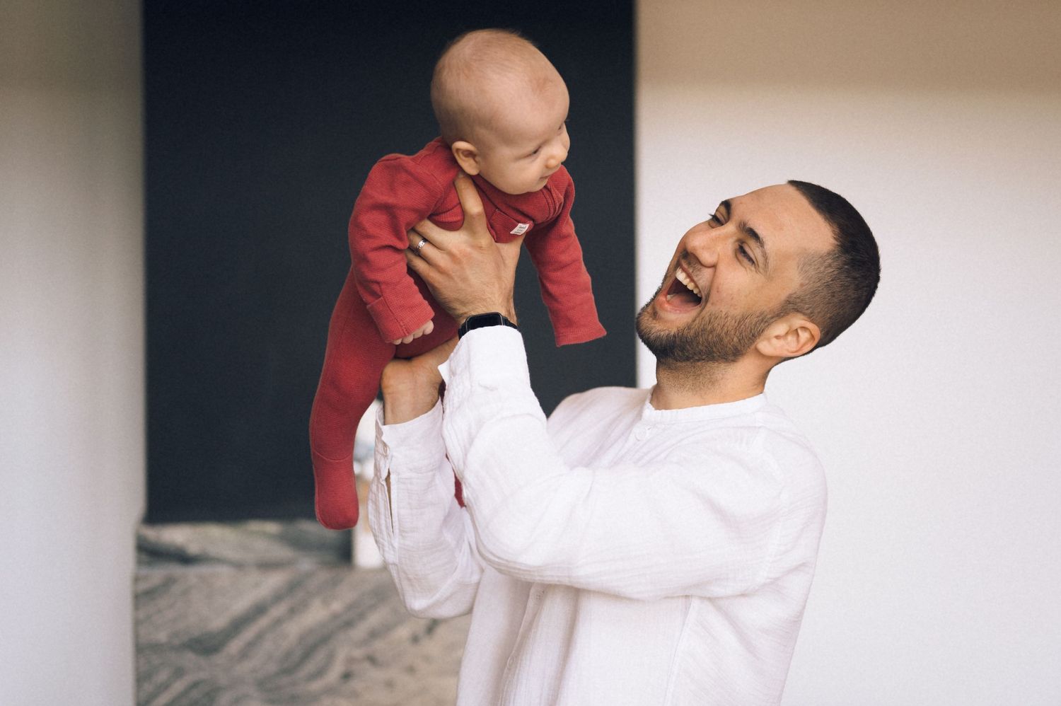 Smiling father in white shirt playfully lifting happy baby in red outfit in bright indoor setting.
