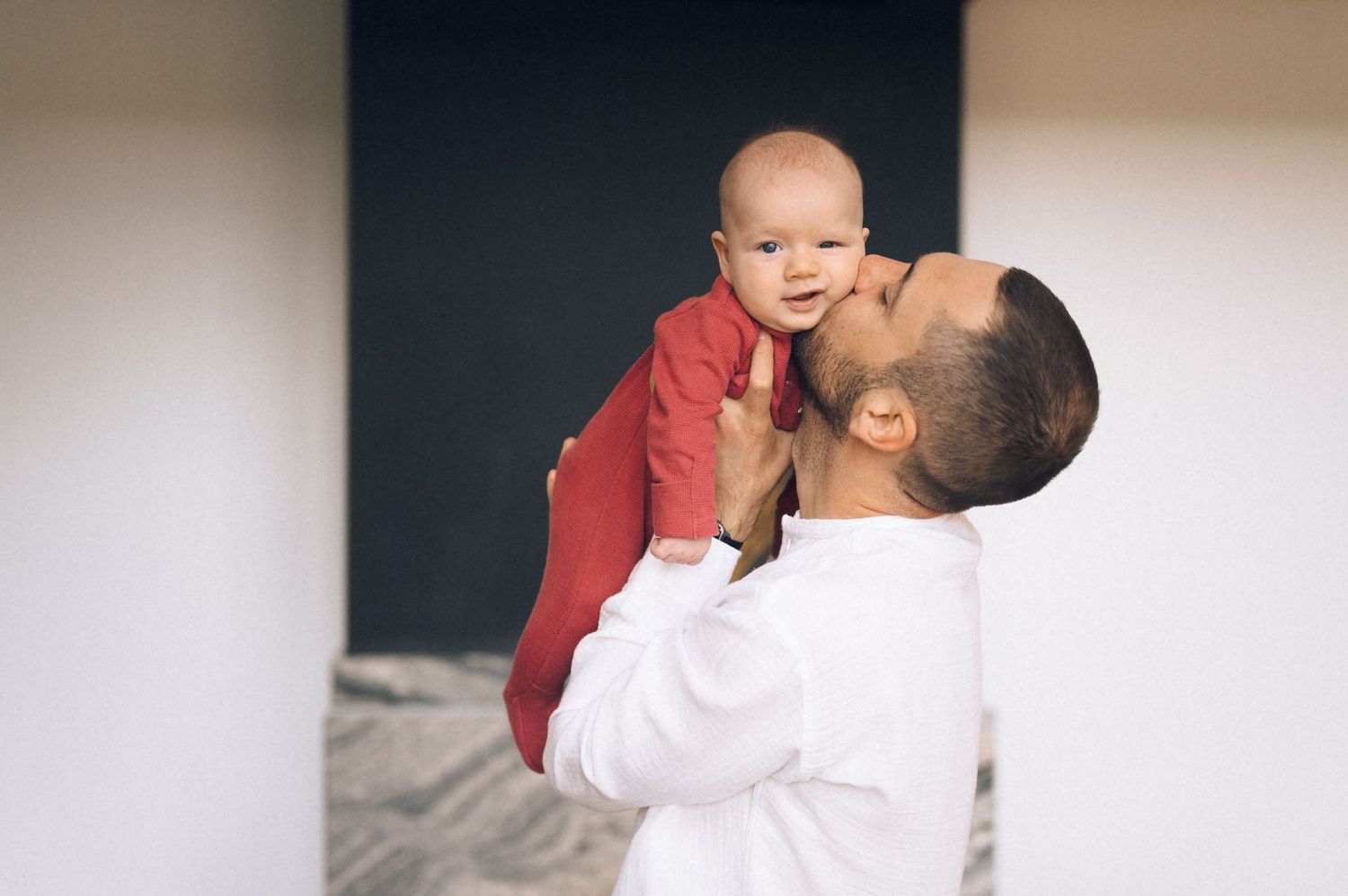 Father in white shirt kissing his baby dressed in red while holding them up.