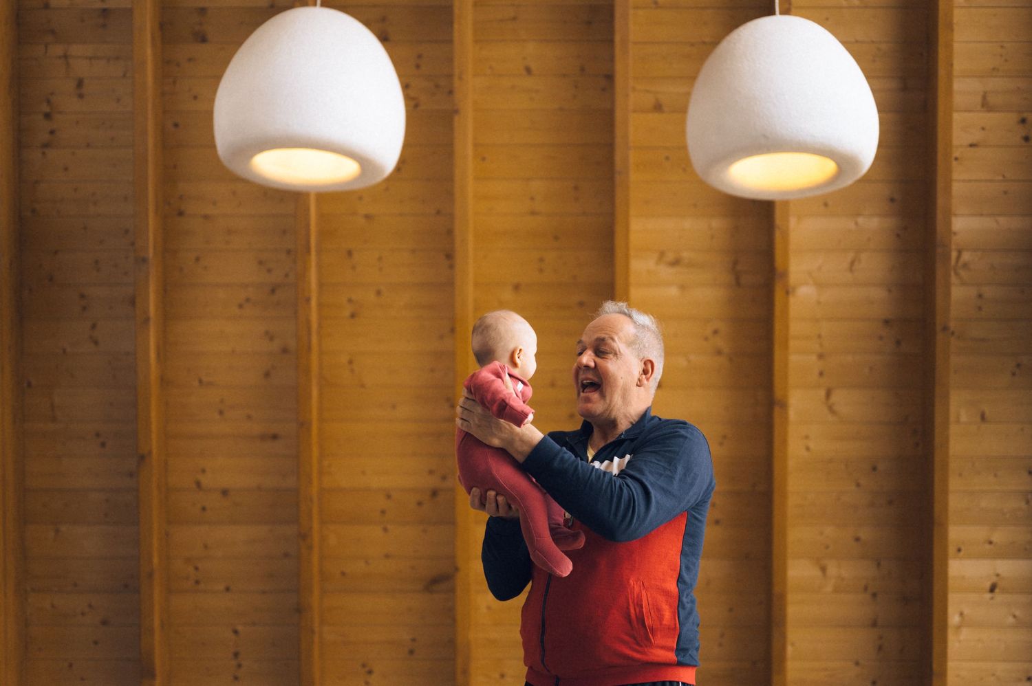 Elderly man joyfully lifting a baby near two white pendant lights against a wooden wall backdrop.