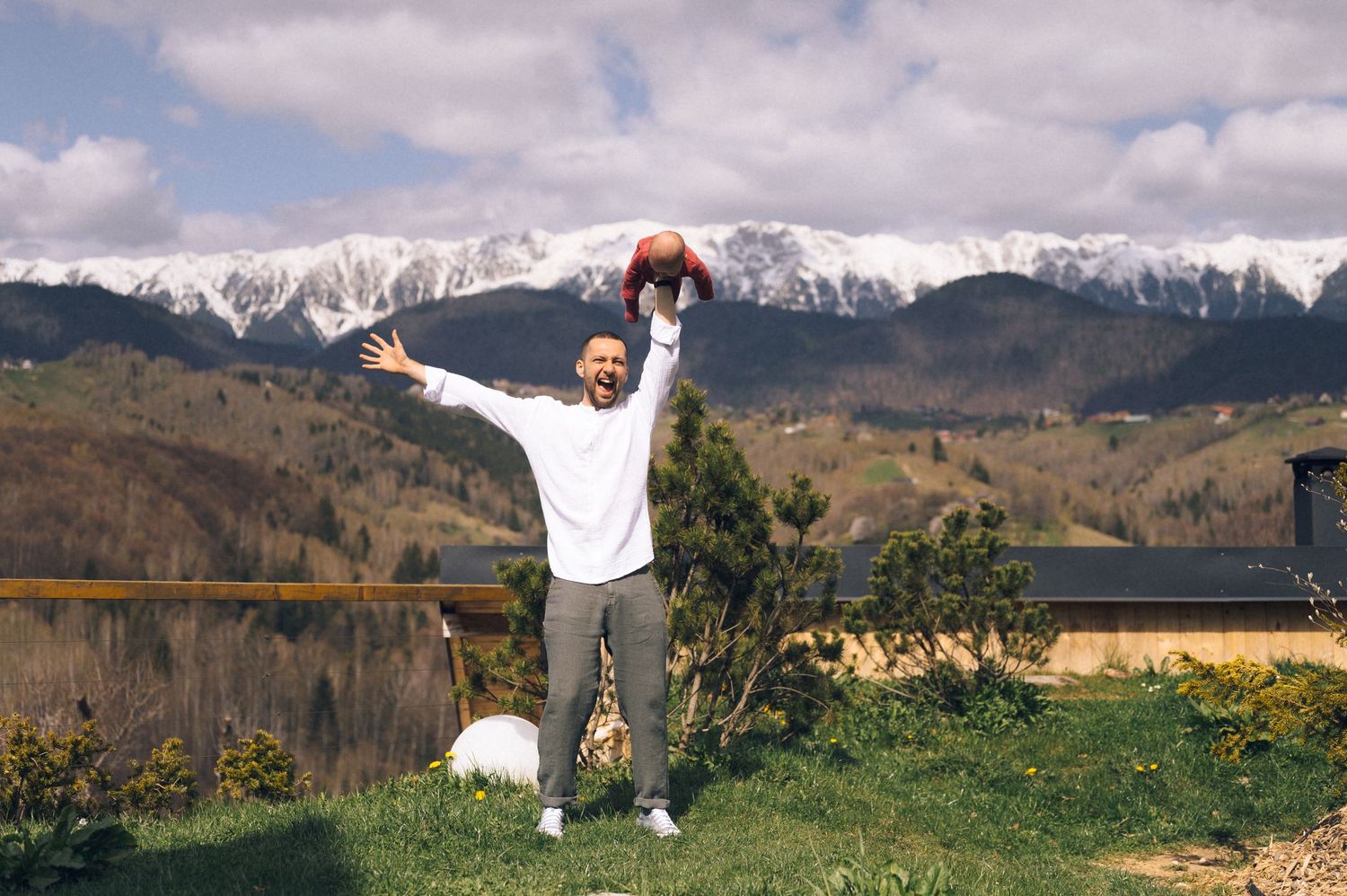 Man celebrating outdoors with arms raised, holding a football, with snow-capped mountains and green landscape in background.