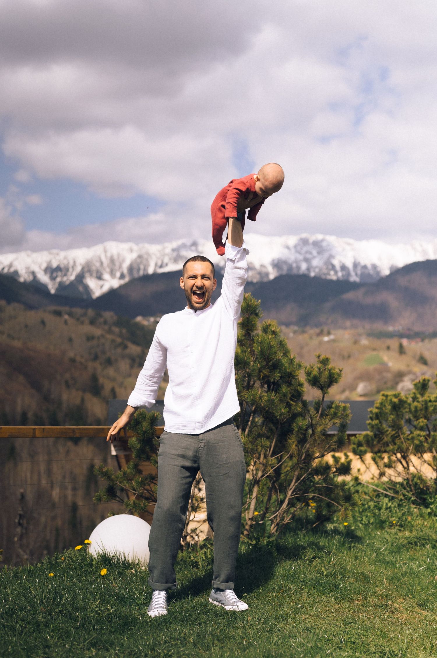 Father joyfully lifting baby in red outfit above his head outdoors with snow-capped mountains in background.