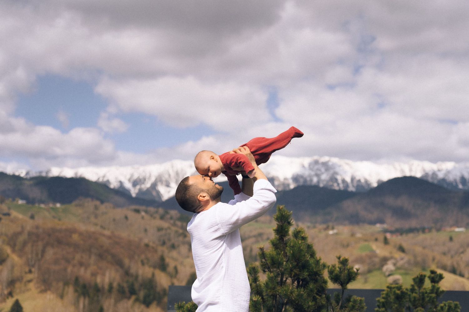 Father joyfully lifting baby in red outfit with snow-capped mountains and cloudy sky in background.