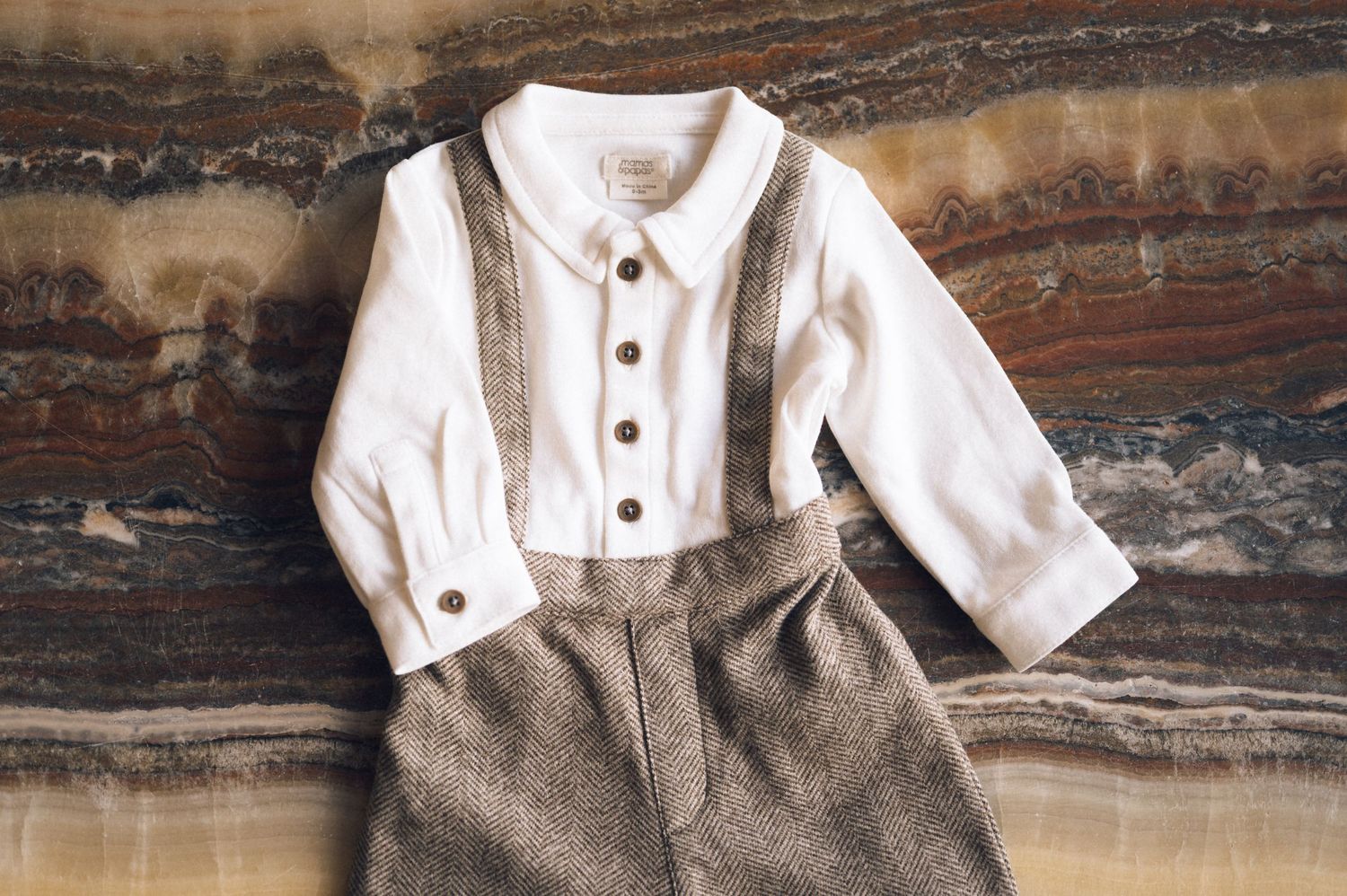 Baby boy outfit with white long-sleeve shirt and brown suspender shorts laid flat on marble surface.