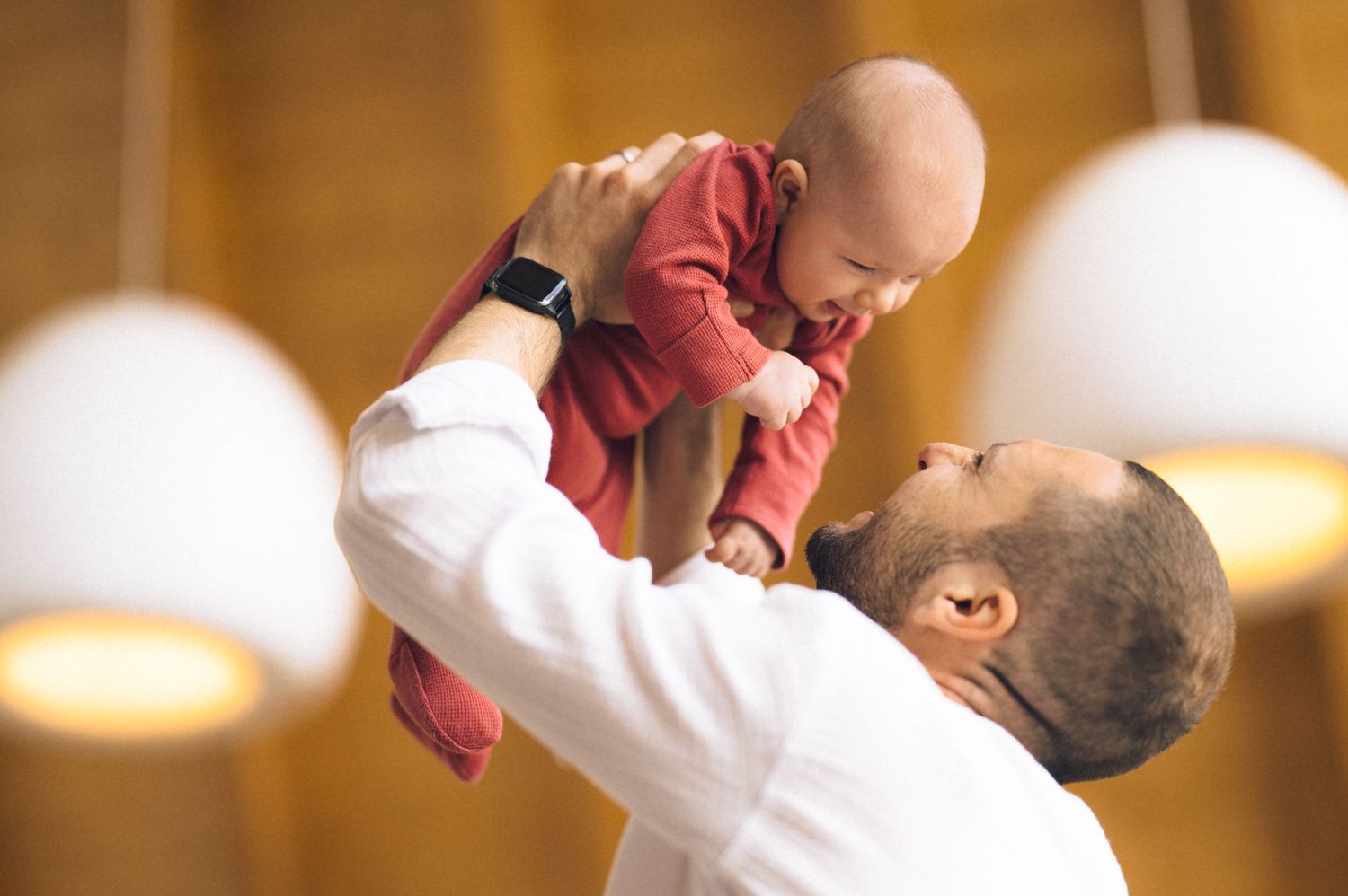 Father joyfully lifting smiling baby in red outfit overhead indoors with warm pendant lights in background.