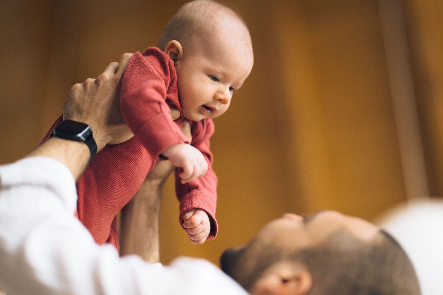 Father joyfully lifting smiling baby in red onesie above his head, sharing a tender bonding moment.