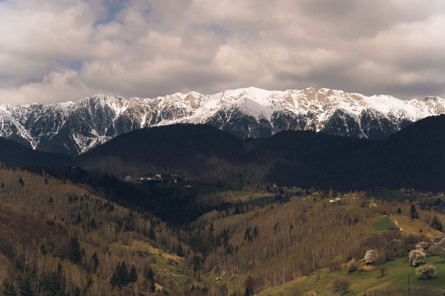 Snow-capped mountain range with forested valleys and small village below under dramatic cloudy sky.