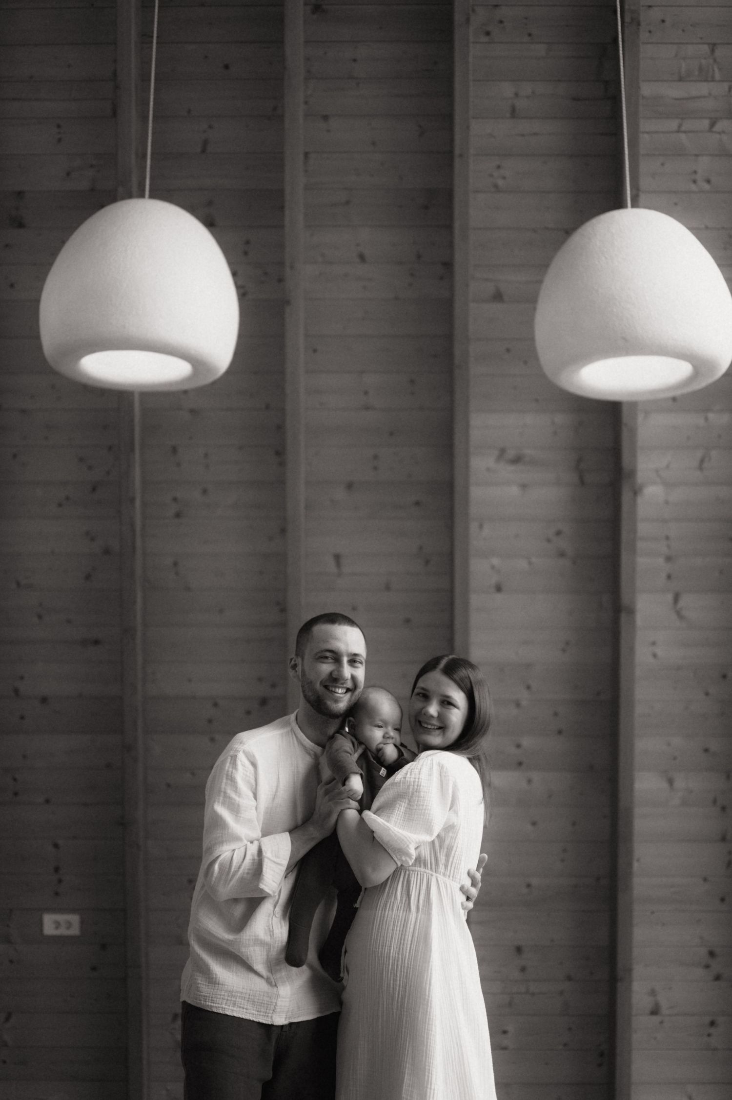 Black and white portrait of smiling couple holding newborn baby under pendant lights against wooden wall.