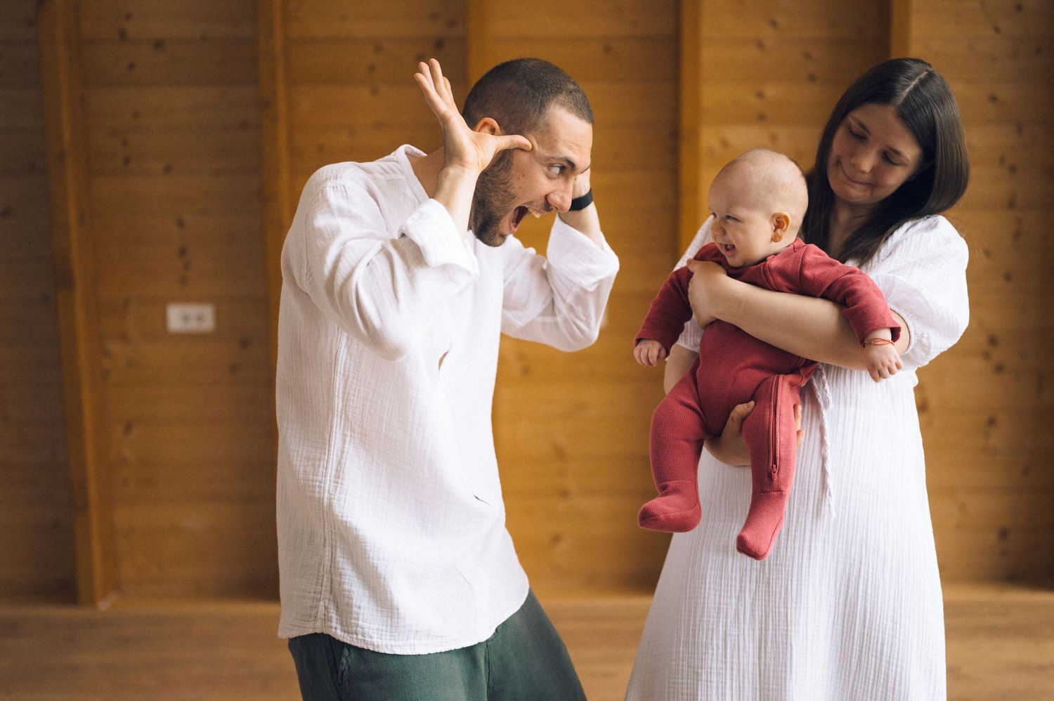 A man and woman in white clothing hold a baby in red outfit inside a wooden room.