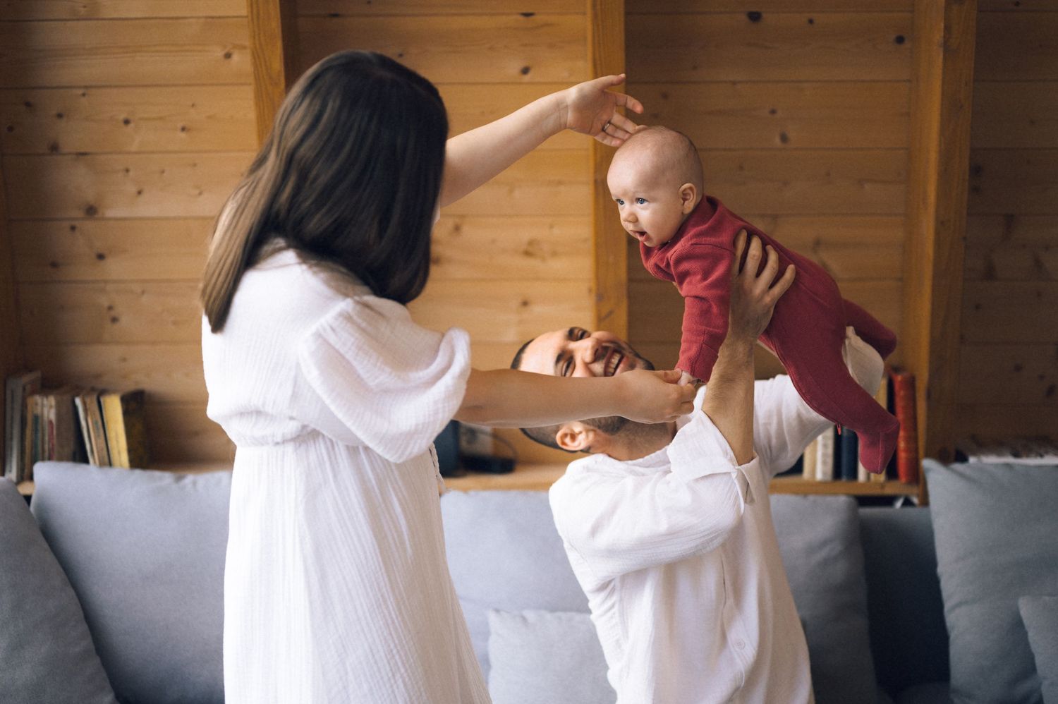 Two women in white dresses playing with a baby in a cozy wooden living room.