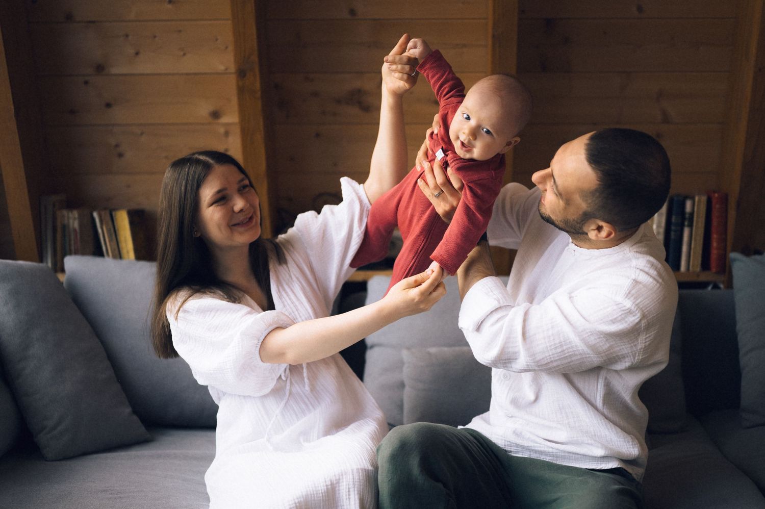 Happy young parents lifting their smiling baby in red outfit while sitting on gray sofa at home.