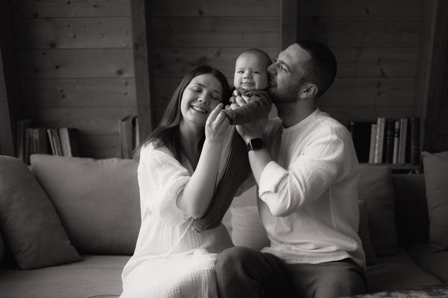 Black and white photo of happy parents holding their baby on a couch in a cozy home setting.