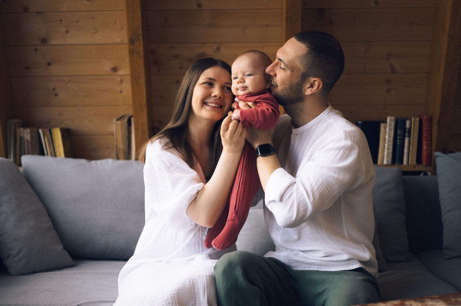Happy young family with baby sitting on sofa at home, parents smiling and kissing newborn infant.