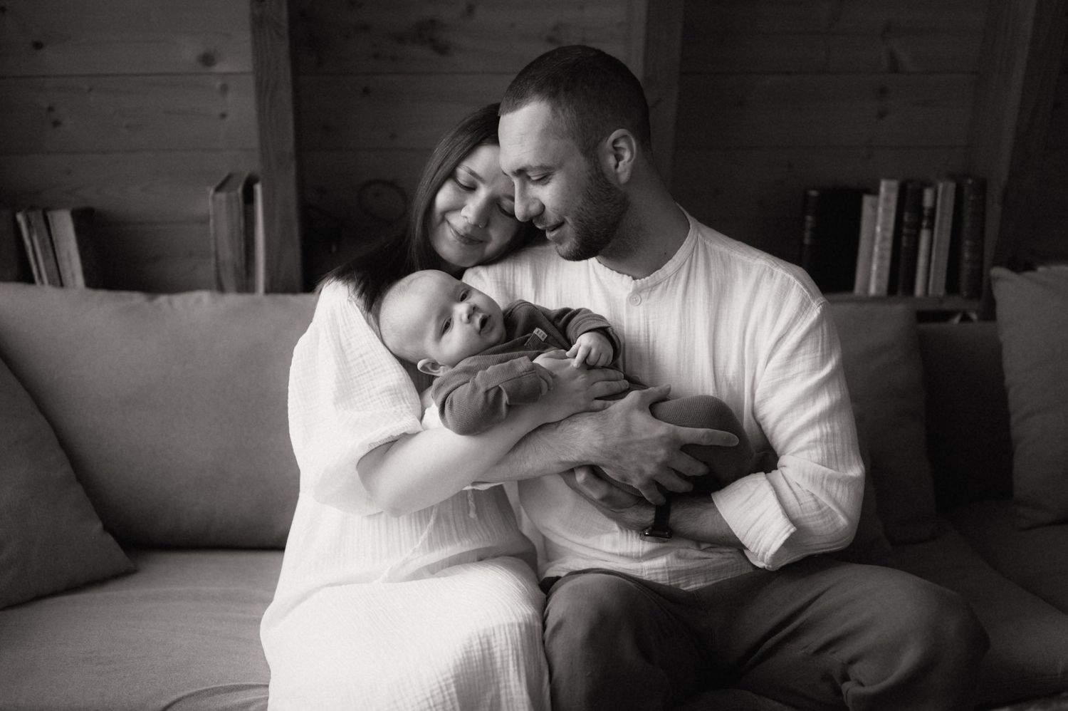 Black and white photo of happy parents sitting on a couch, lovingly cradling their newborn baby together.