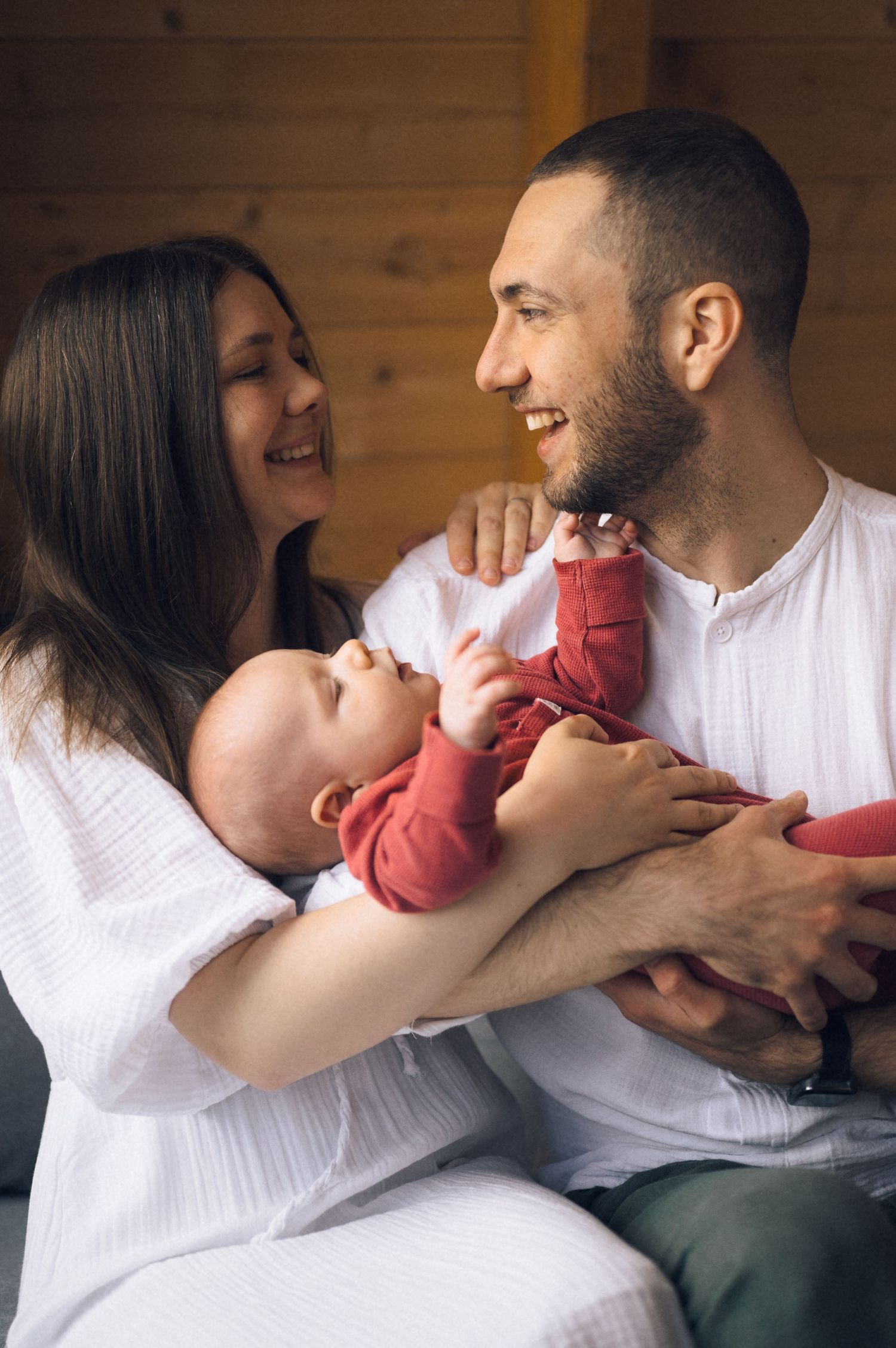 Happy young parents smiling at each other while holding their newborn baby dressed in red at home.