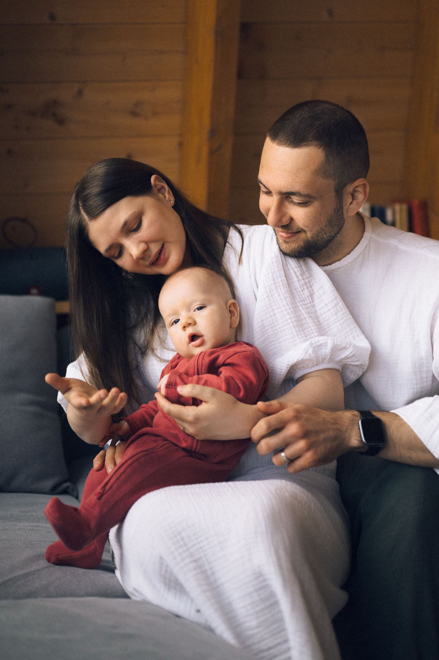 Happy young family with baby sitting on couch, parents in white shirts smiling at infant in red outfit.