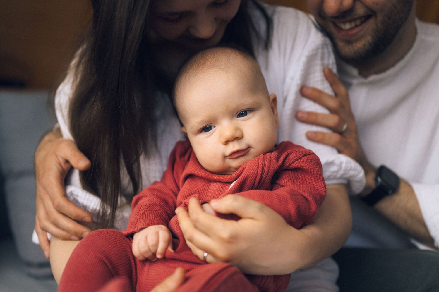Happy young parents holding their smiling baby dressed in a red onesie, sharing a tender family moment together.