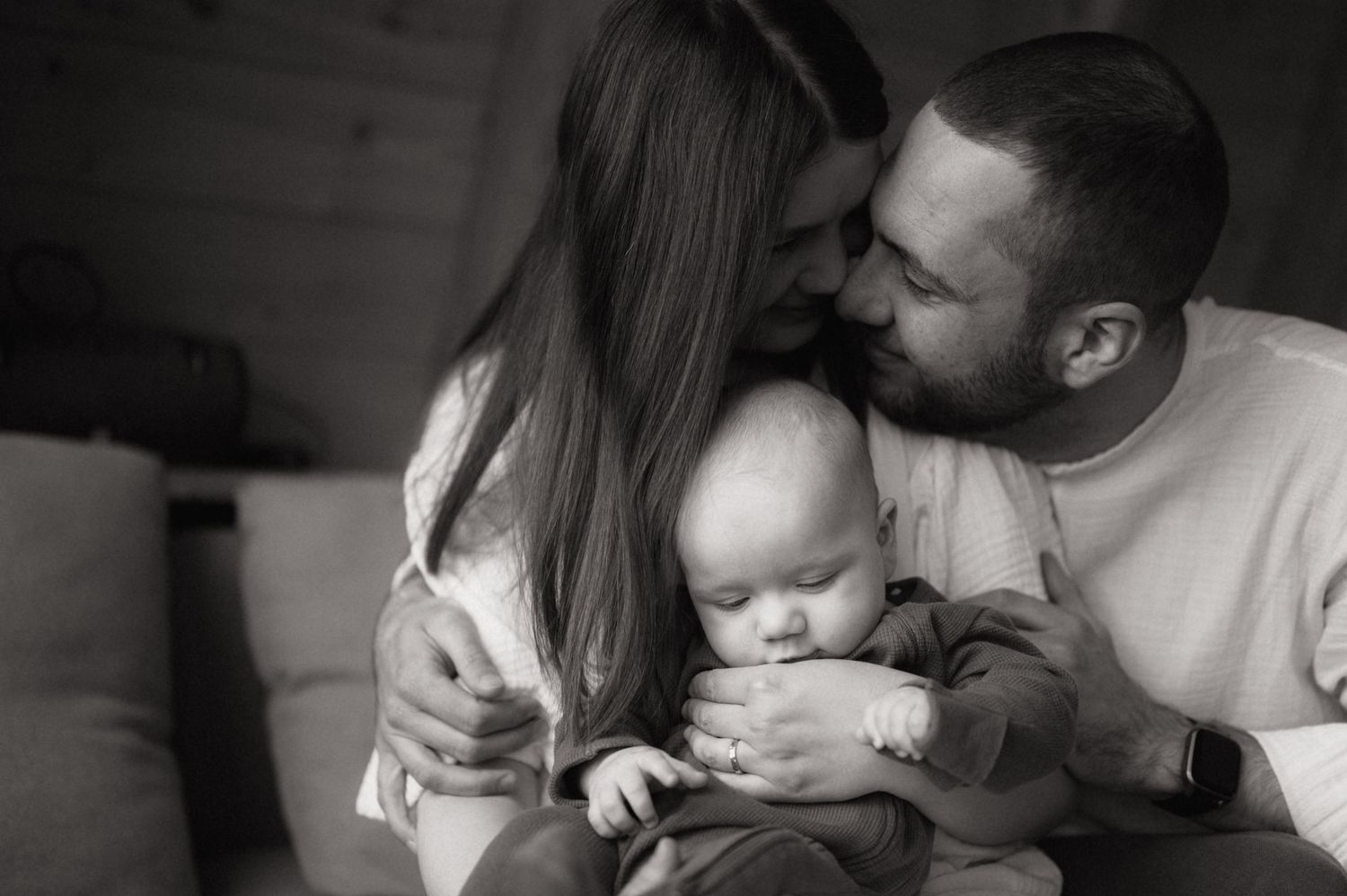 Black and white photo of parents kissing their newborn baby in a cozy indoor setting.