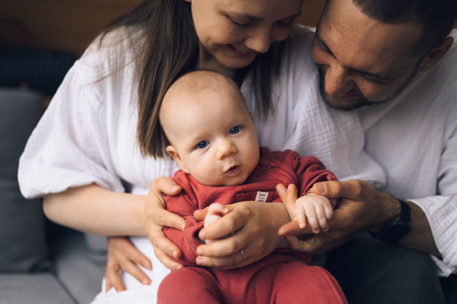 Parents lovingly holding their alert newborn baby dressed in a red onesie during a family portrait session.