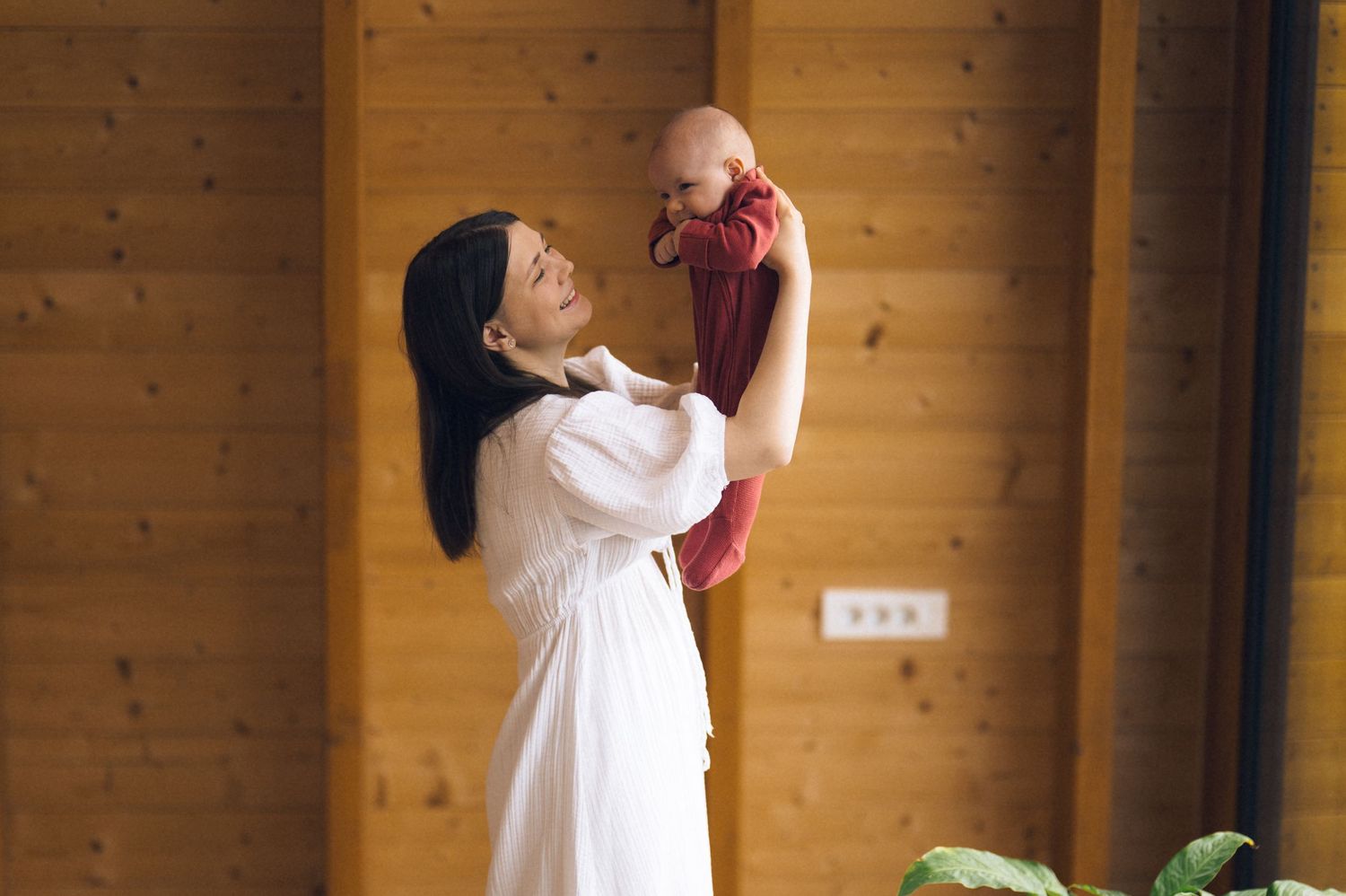 A mother in a white dress joyfully lifts her baby in red clothing indoors with wooden walls.