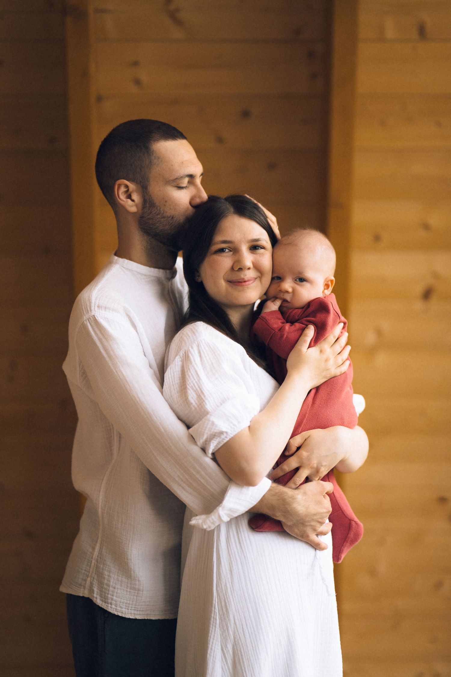 Young family portrait with father embracing mother holding newborn baby wrapped in red blanket indoors.