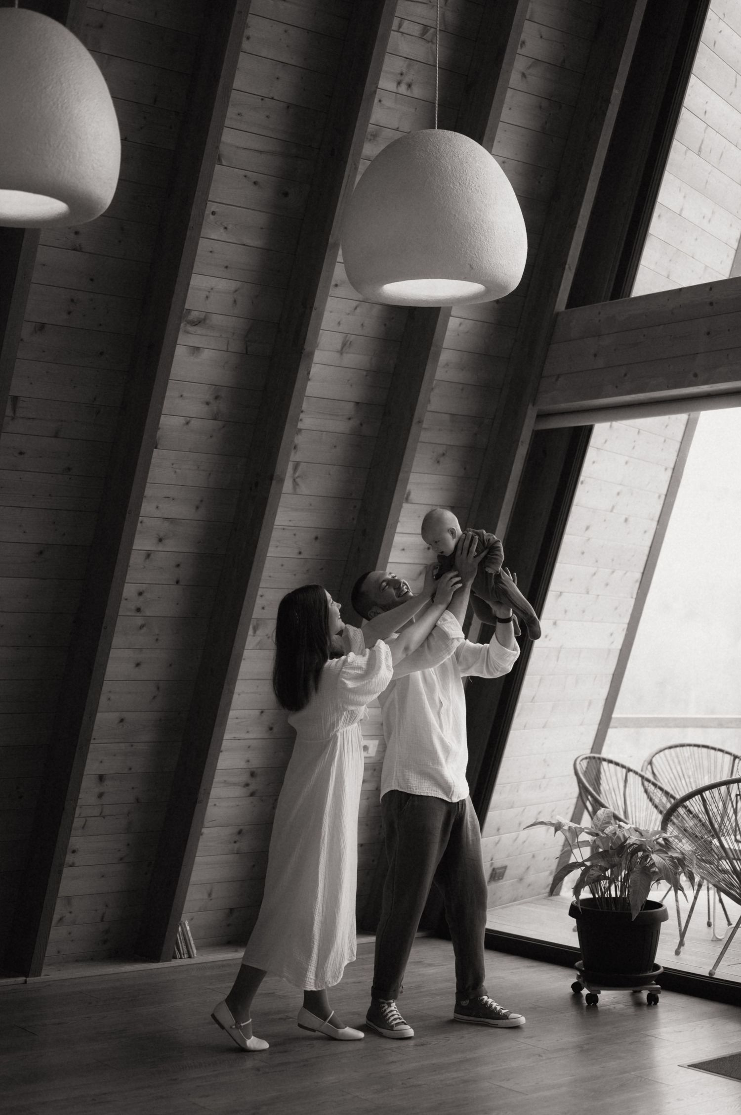 Black and white photo of a couple lifting their baby in a modern wooden interior with pendant lights.