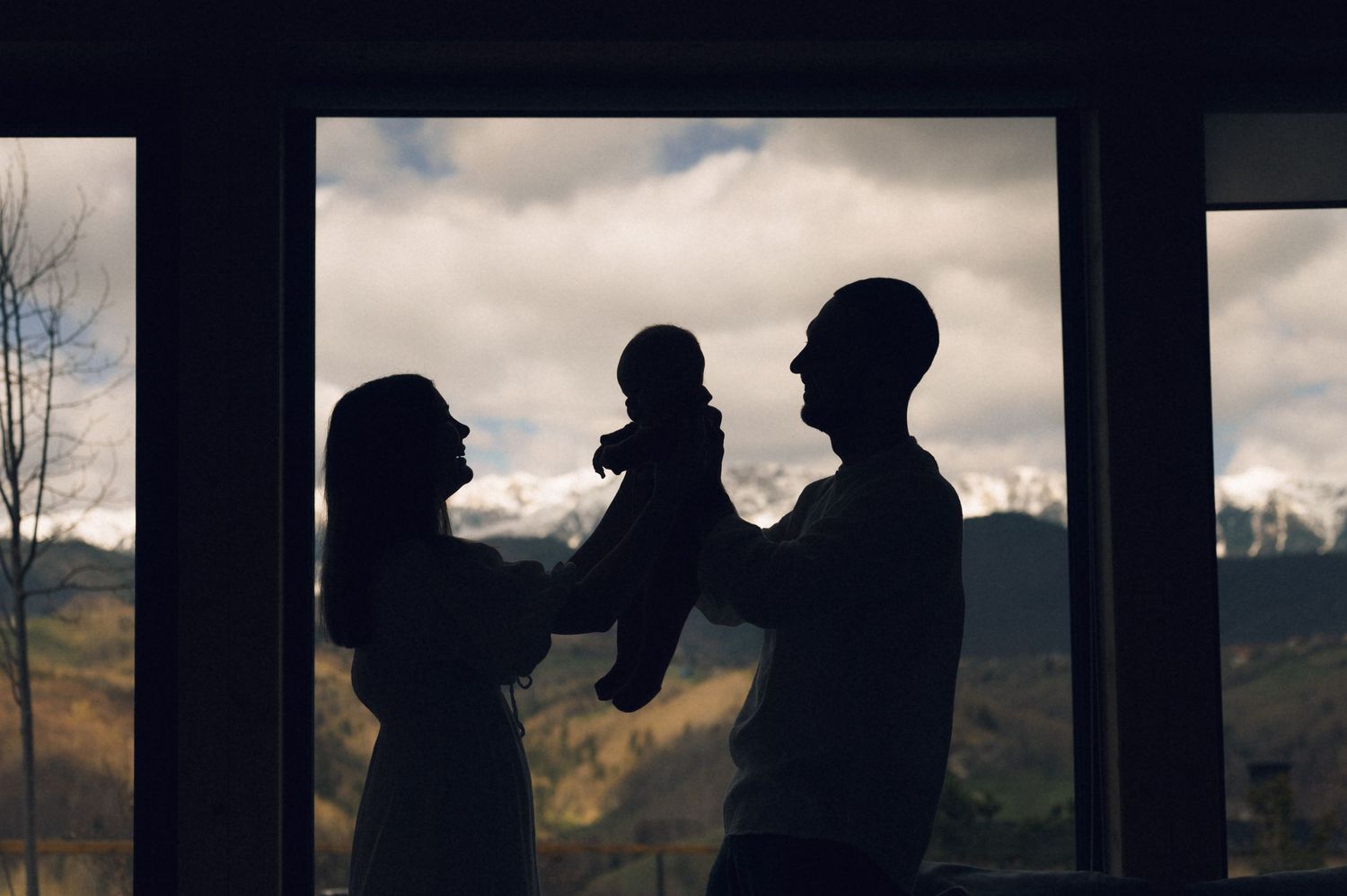 Silhouette of parents holding baby against mountain window backdrop.