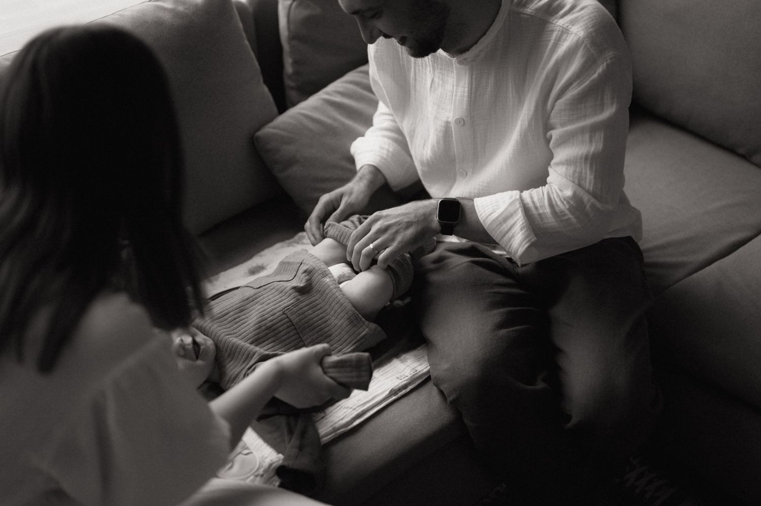 Black and white photo of a father cradling his newborn baby on a couch with a woman nearby.