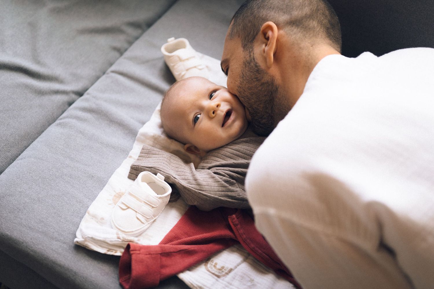 Father smiling and bonding with happy baby lying on gray sofa, tender parenting moment.