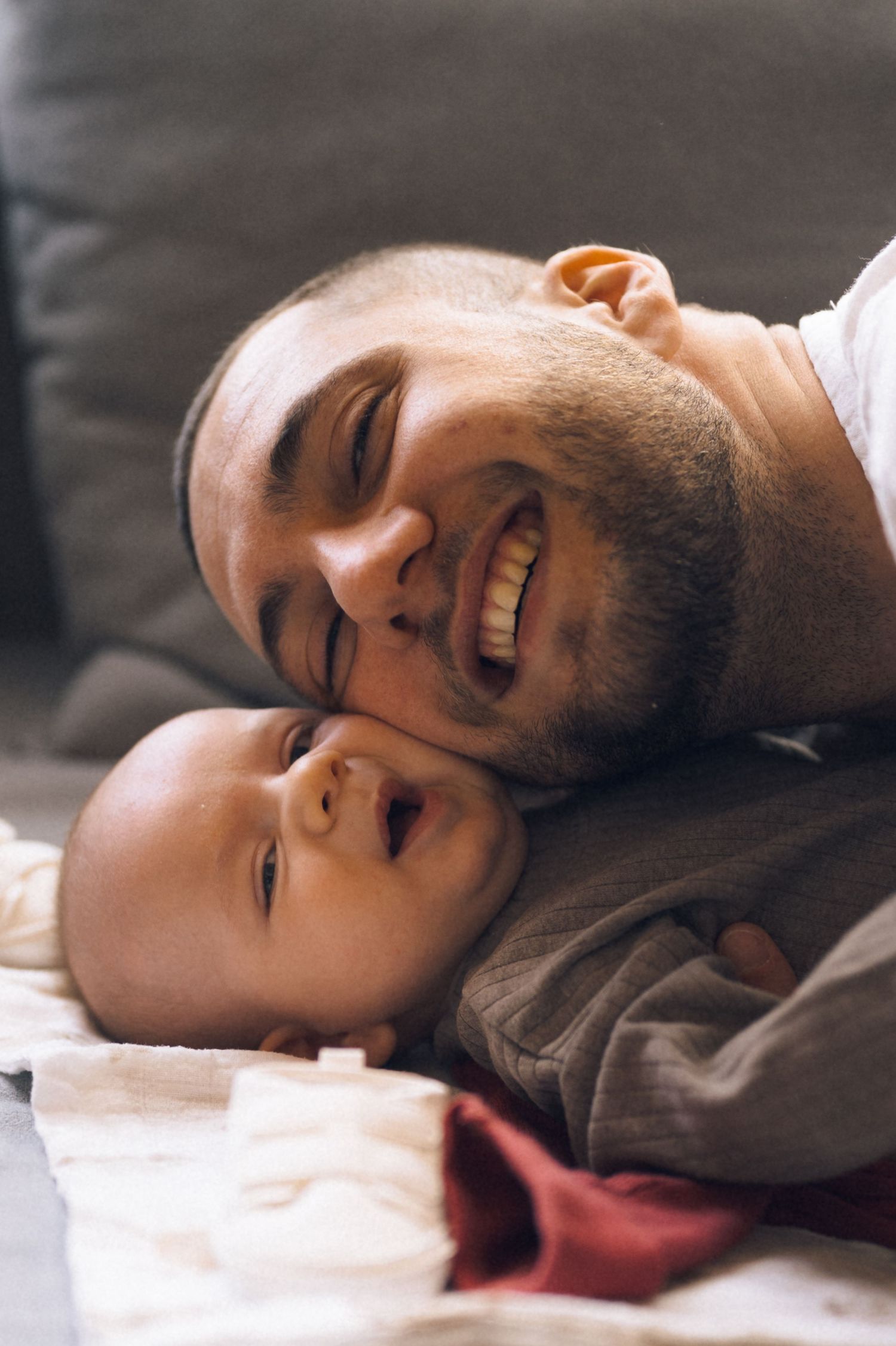 Smiling father lying down cheek-to-cheek with laughing baby on a blanket near a gray couch.