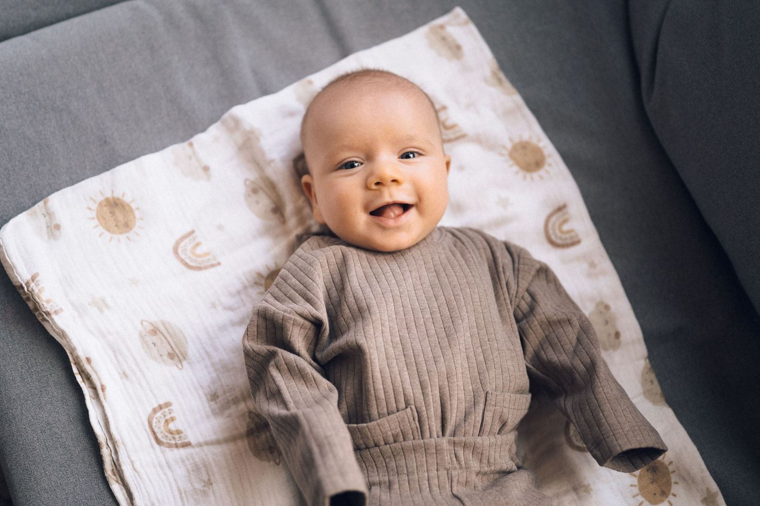 Smiling baby in brown ribbed outfit lying on celestial print blanket on gray sofa.