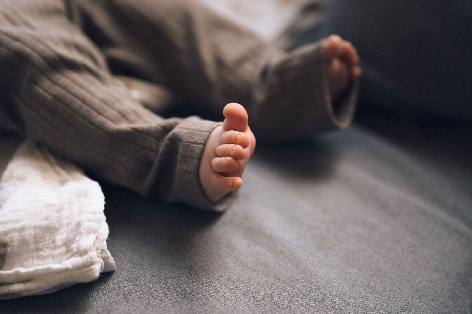 Close-up of a newborn baby's tiny foot in a knit onesie on a gray surface.