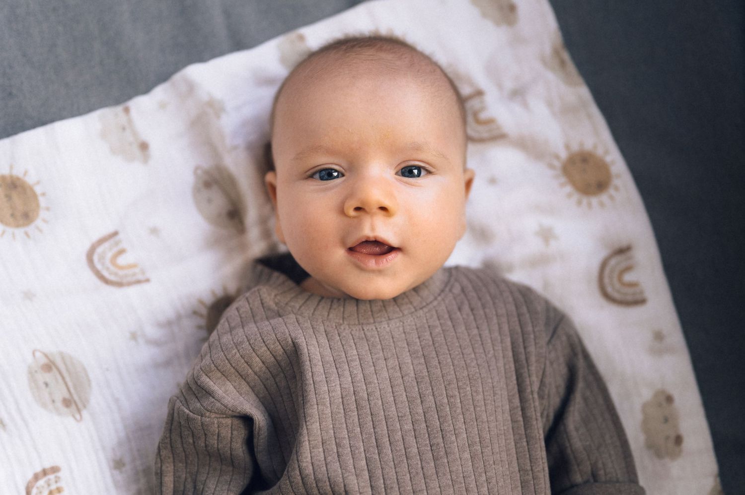 Adorable newborn baby in gray ribbed outfit lying on a sun and rainbow patterned blanket, looking up curiously.