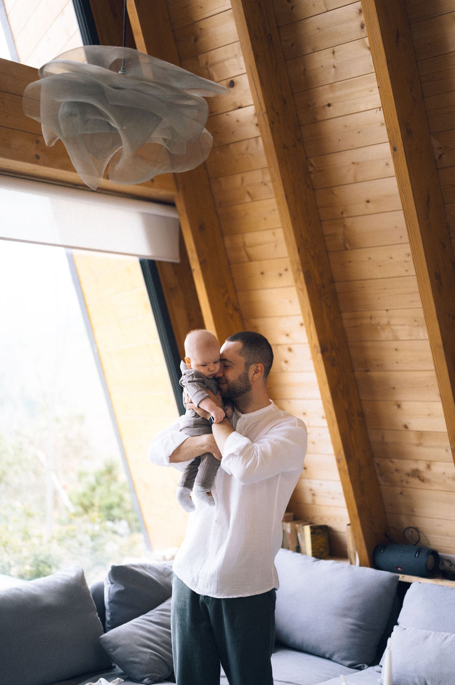 Father kissing his baby in a modern wooden cabin living room with large windows and gray sofas.