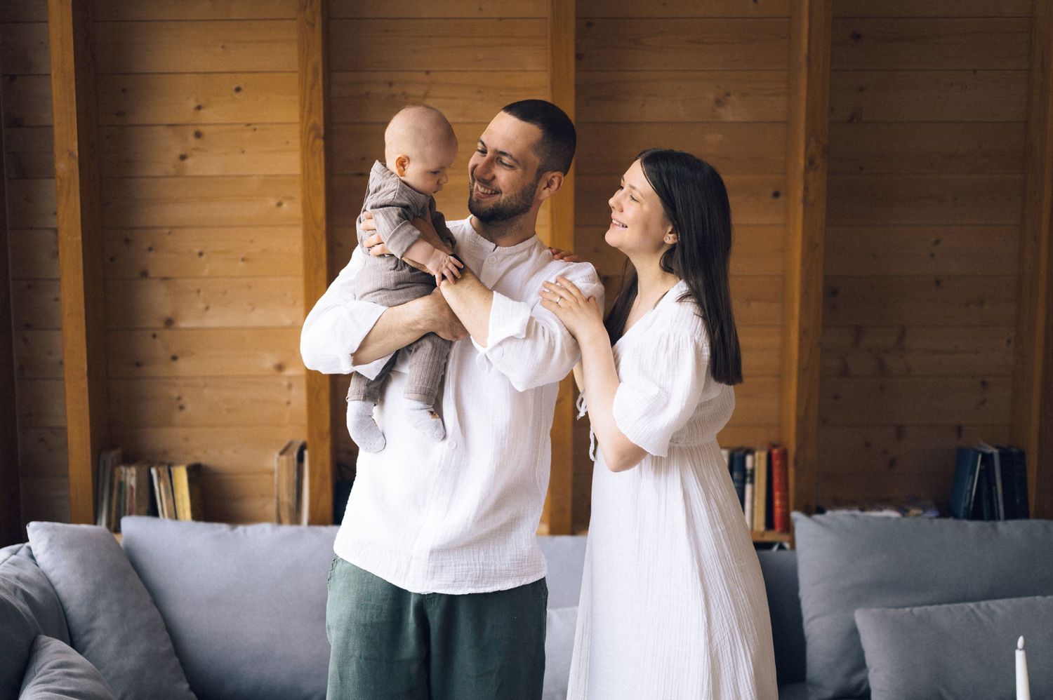 Happy young family with baby in cozy wooden living room, parents smiling and holding infant together.