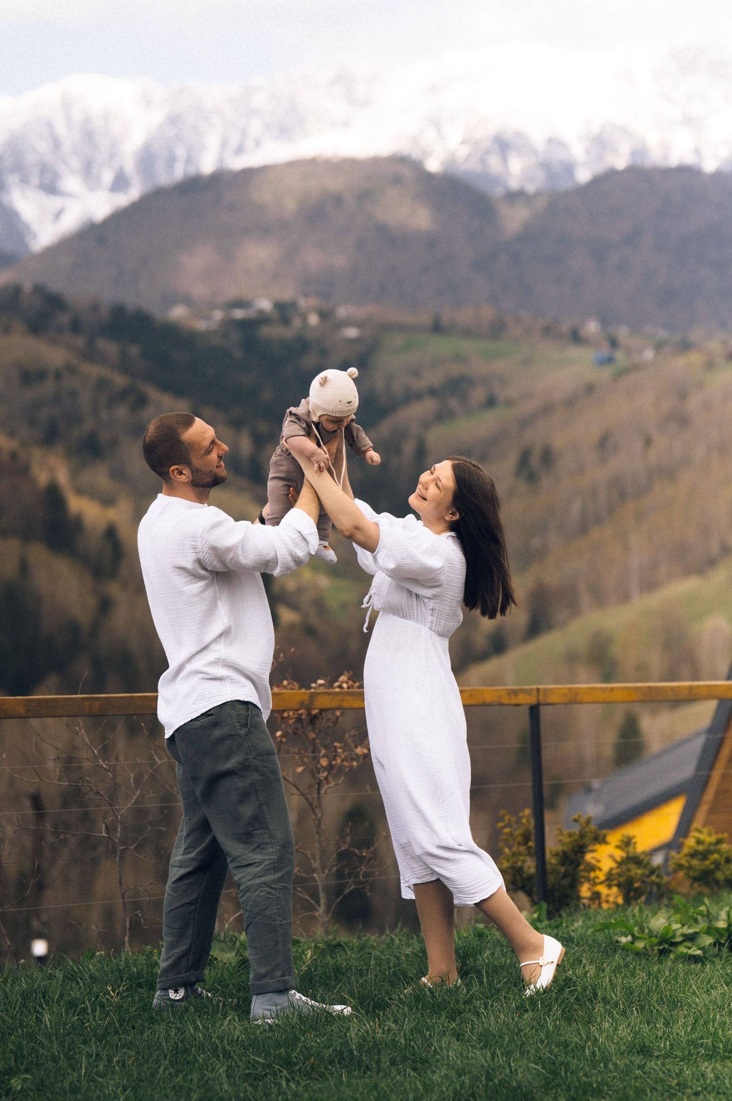 Happy couple lifting their baby outdoors with snow-capped mountains and green hills in the background.
